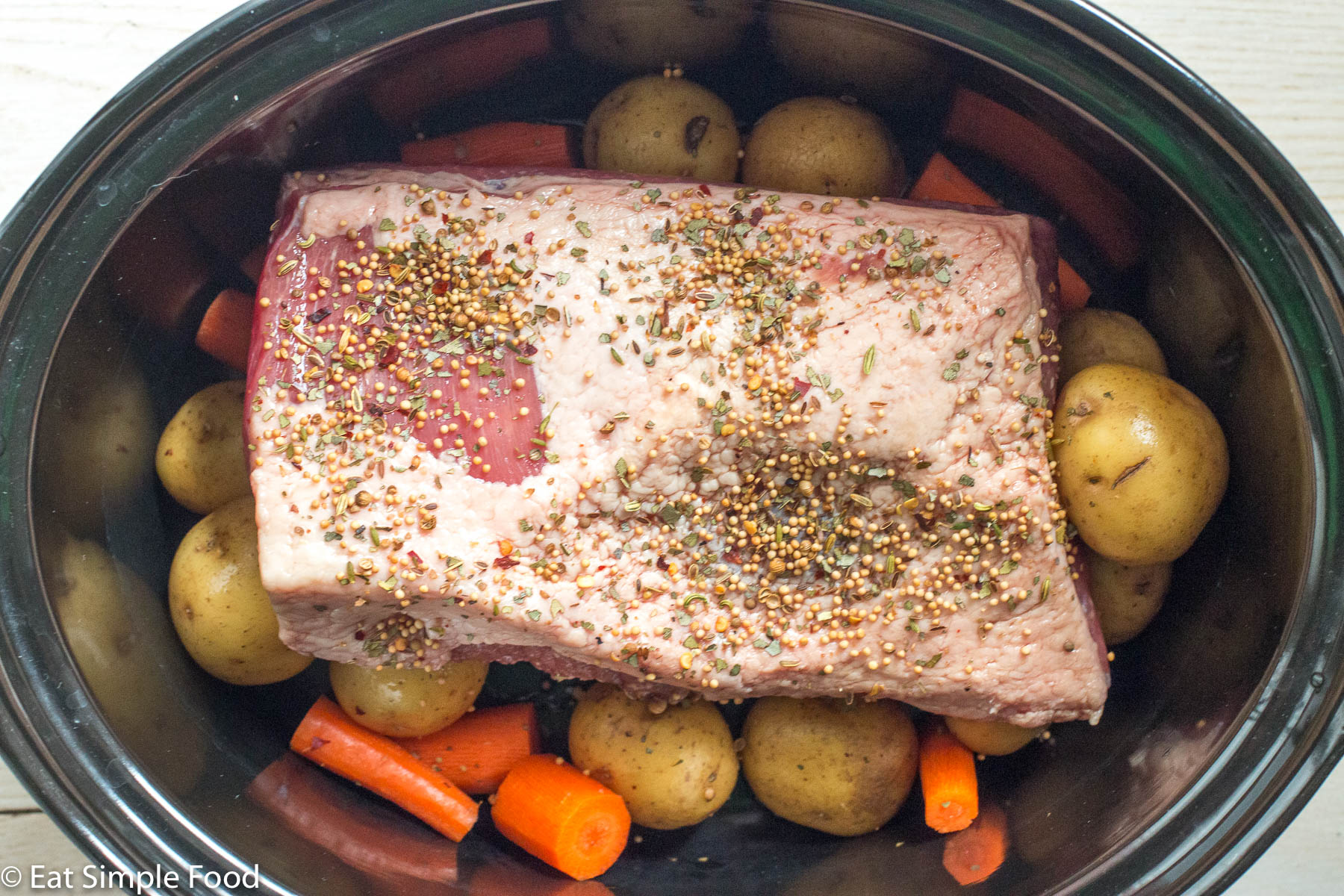 Top down view of a raw fat side up beef brisket with spices in a crockpot sitting on top of baby potatoes and chunks of carrots.