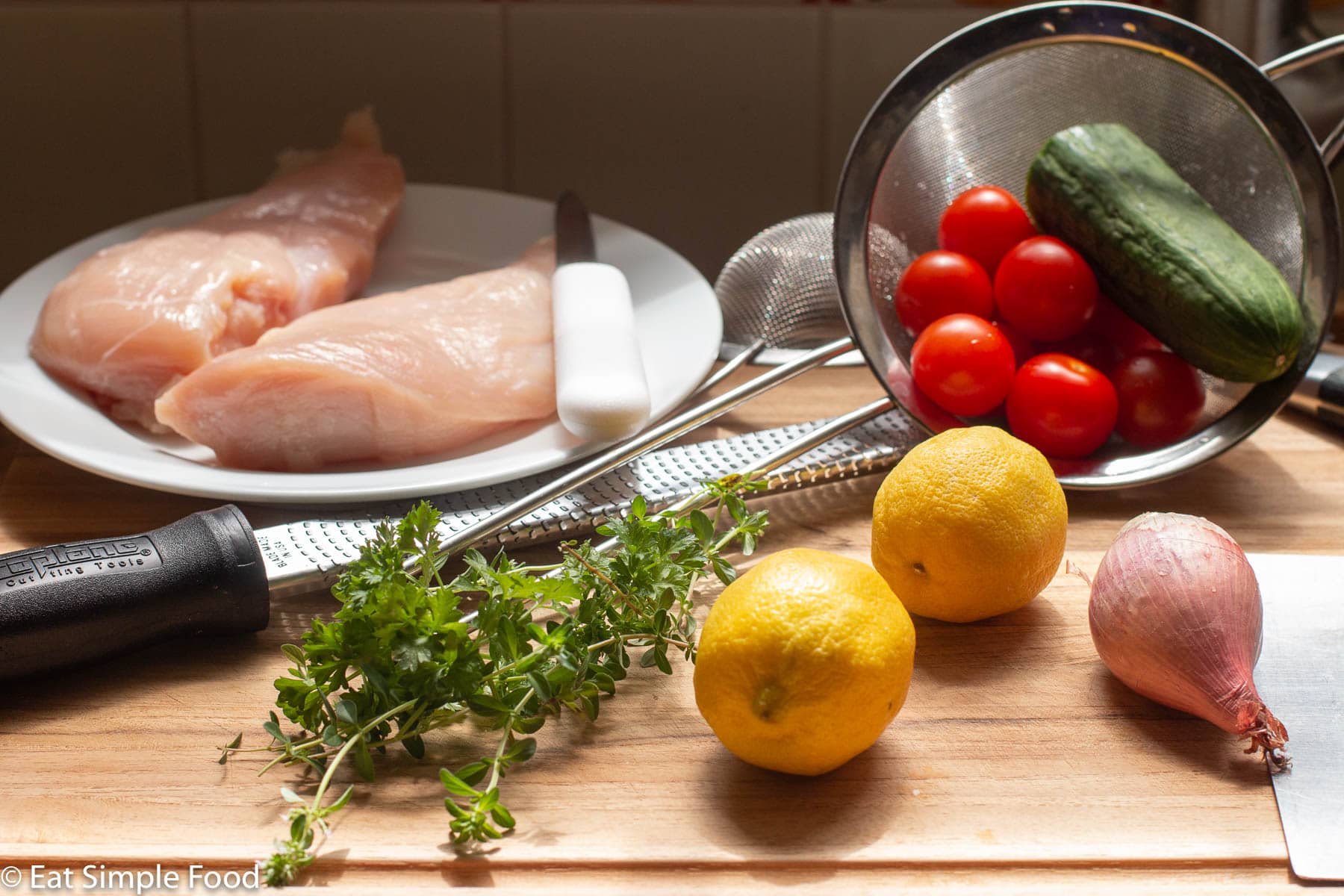 Raw ingredients on a cutting board: 2 lemons, 1 shallot, fresh parsley and thyme, strainer of cherry tomatoes and ½ a cucumber, plate with two chicken breasts.
