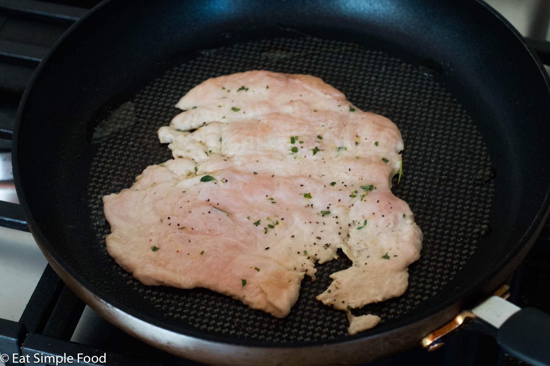Raw flattened chicken breast cooking in a black skillet. Side view.