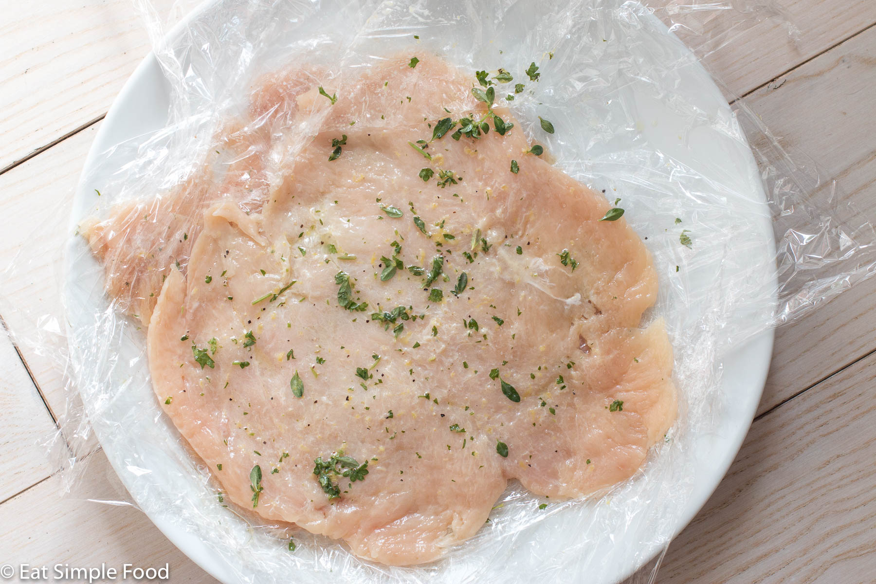 Flattened pieces of chicken stacked on a white plate with saran wrap in between each piece of chicken. Lemon zest, olive oil, and fresh herbs sprinkled on top.