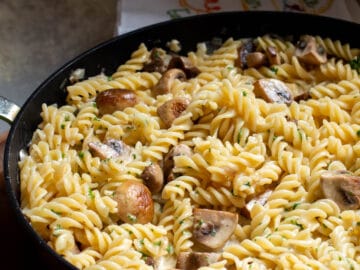 Close up of pan of fusilli pasta with quartered mushrooms in a light cream sauce with parsley garnish.