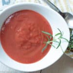 White bowl of plum coulis sauce with a fresh sprig of rosemary on it. Napkin and spoon on the side. Close up.