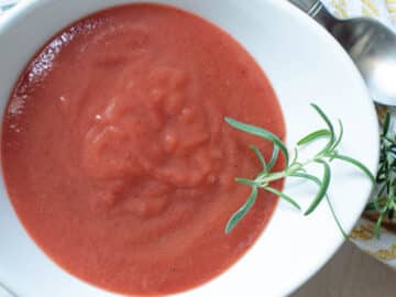 White bowl of plum coulis sauce with a fresh sprig of rosemary on it. Napkin and spoon on the side. Close up.