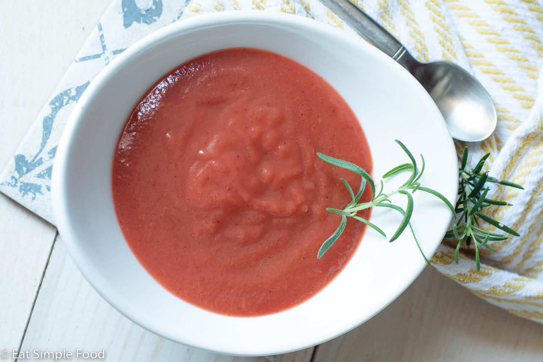 White bowl of plum coulis sauce with a fresh sprig of rosemary on it. Napkin and spoon on the side.