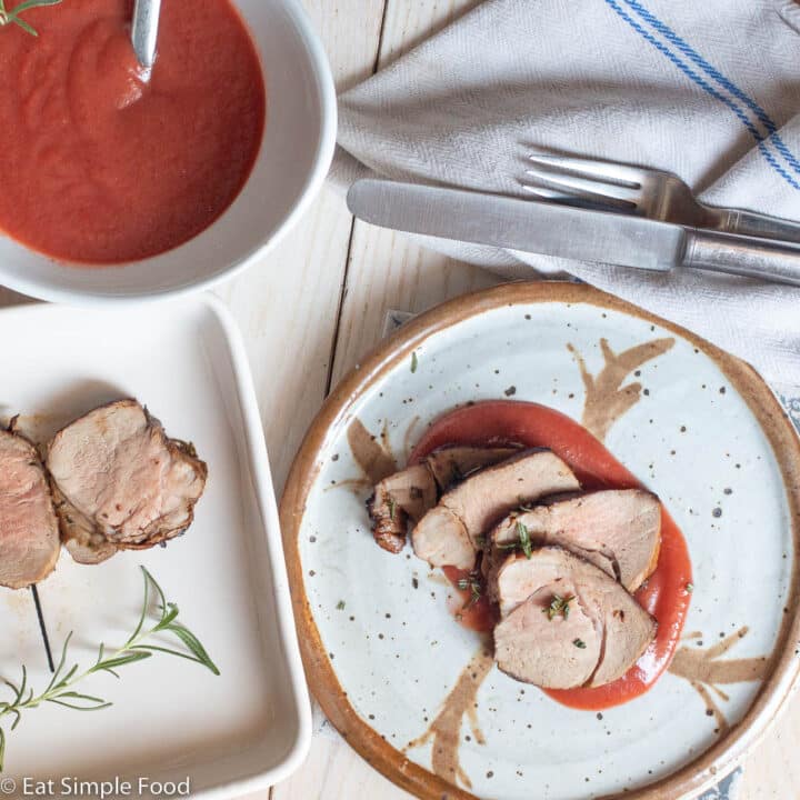 Close up top down view of sliced pork tenderloin on a plate with a plum purple sauce underneath. Bowl of purple sauce and pork tenderloin on the side.