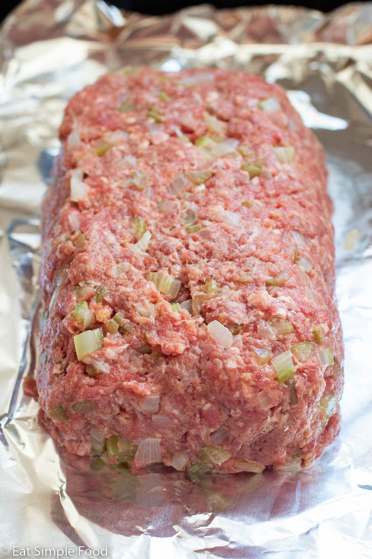 Raw meatloaf with diced celery and onions on an aluminum lined baking sheet.