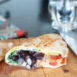 Browned flour tortilla quesadilla cut in half with filling showing on a wood cutting board with a chef's knife. Ingredients are black beans, arugula, cheese, and tomatoes. Close up.