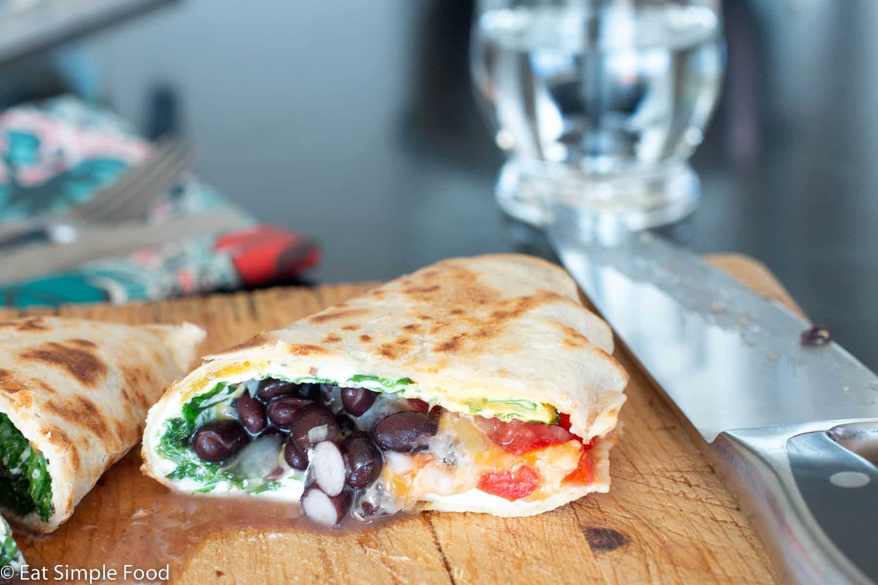 Browned flour tortilla quesadilla cut in half with filling showing on a wood cutting board with a chef's knife. Ingredients are black beans, arugula, cheese, and tomatoes.