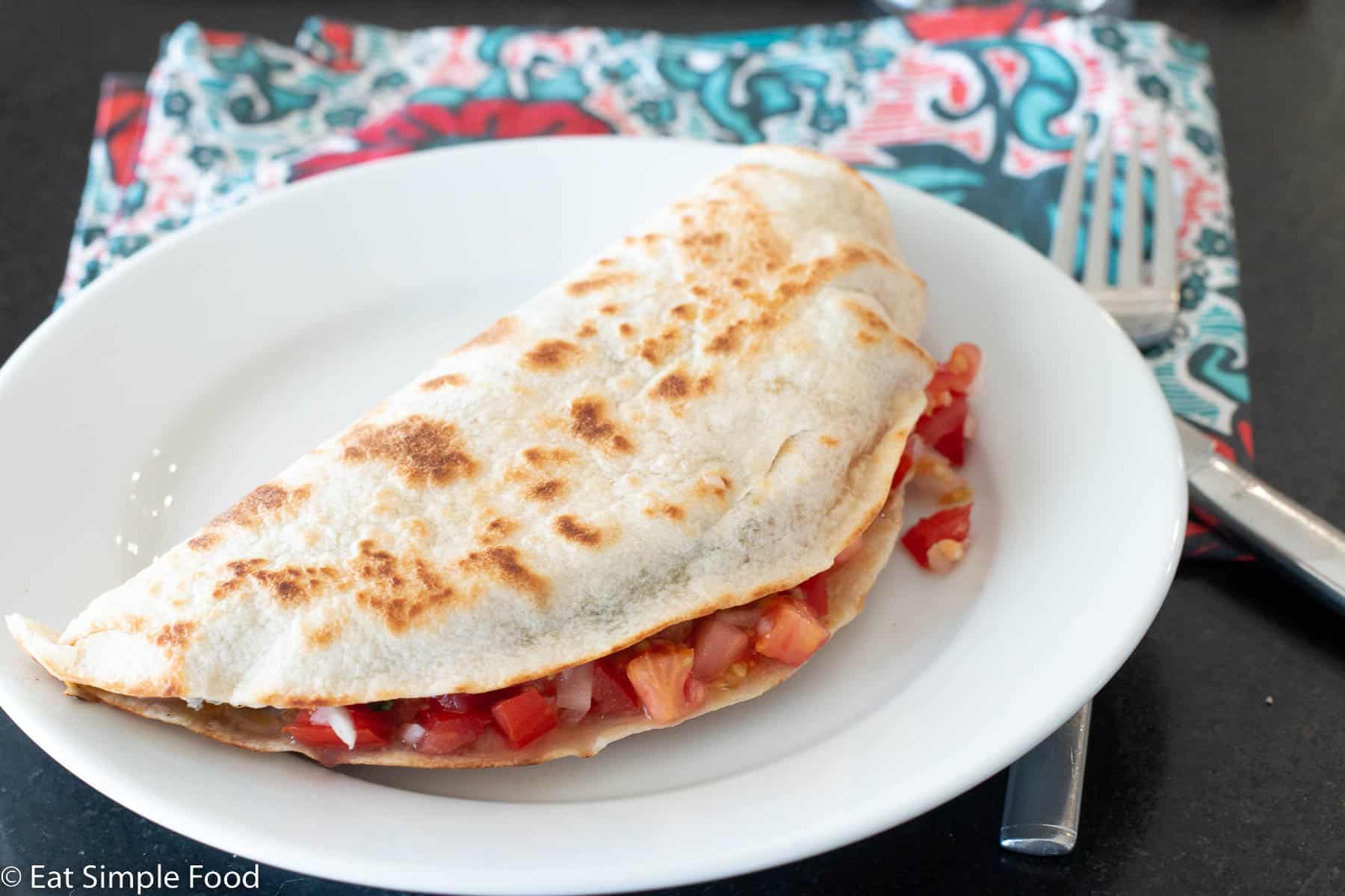 Browned flour tortilla folded in half filled with ingredients like tomatoes on a white plate. Fork and knife and blue and red napkin in background.