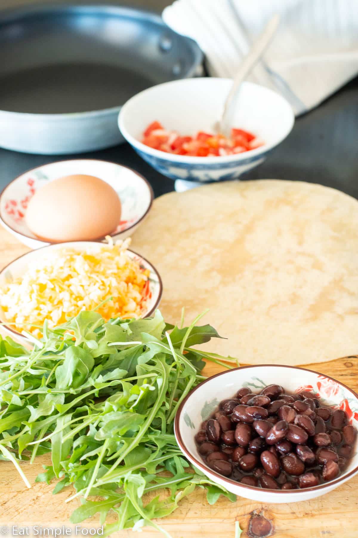Ingredients on a wood cutting board with a nonstick skillet and napkin in background. Ingredients: tomato salsa, whole egg in shell, shredded yellow cheese, handful of arugula, small ramiken of black beans.