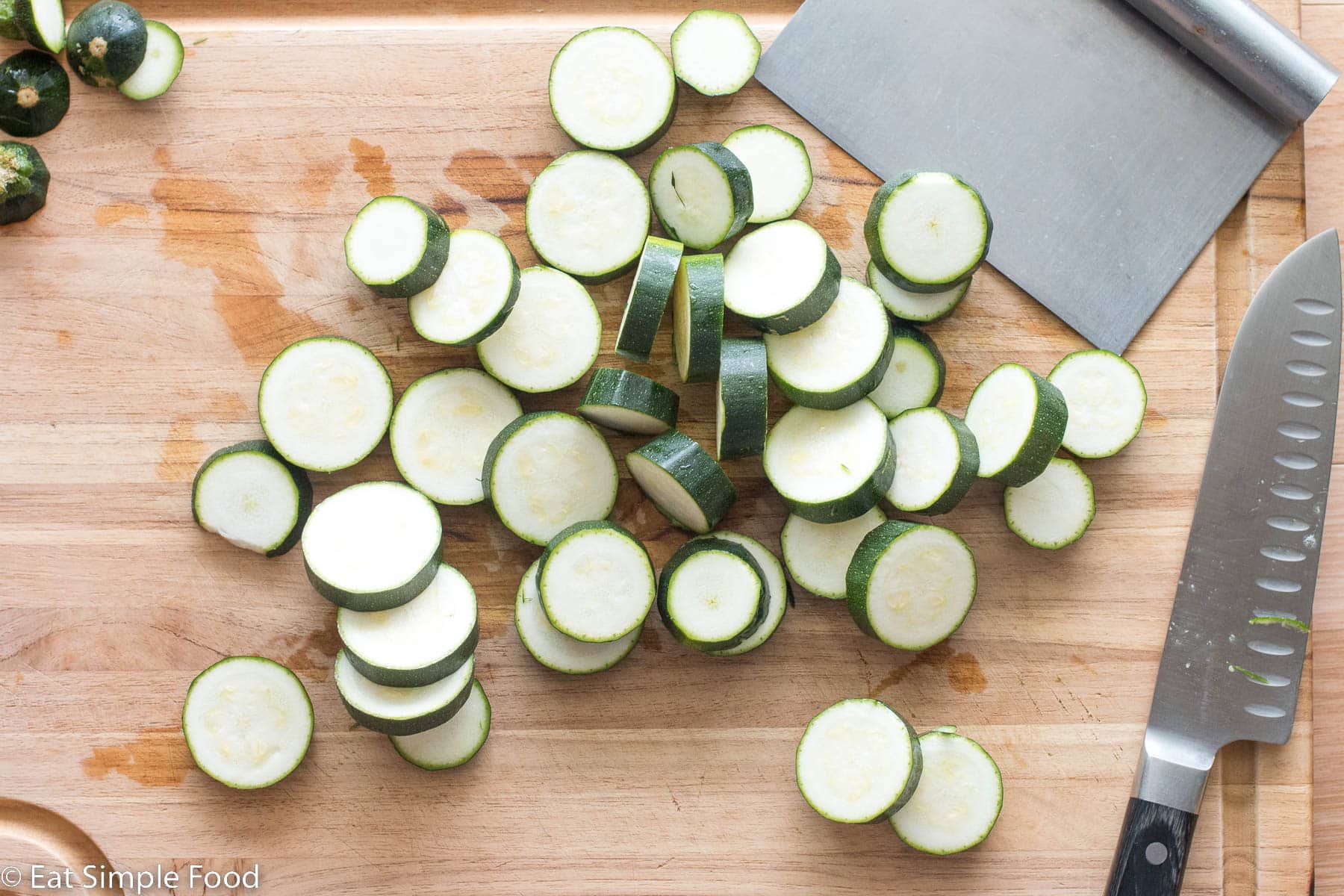Raw zucchini sliced half an inch thick on a wood cutting board. Pastry scrapper and knife on the side.