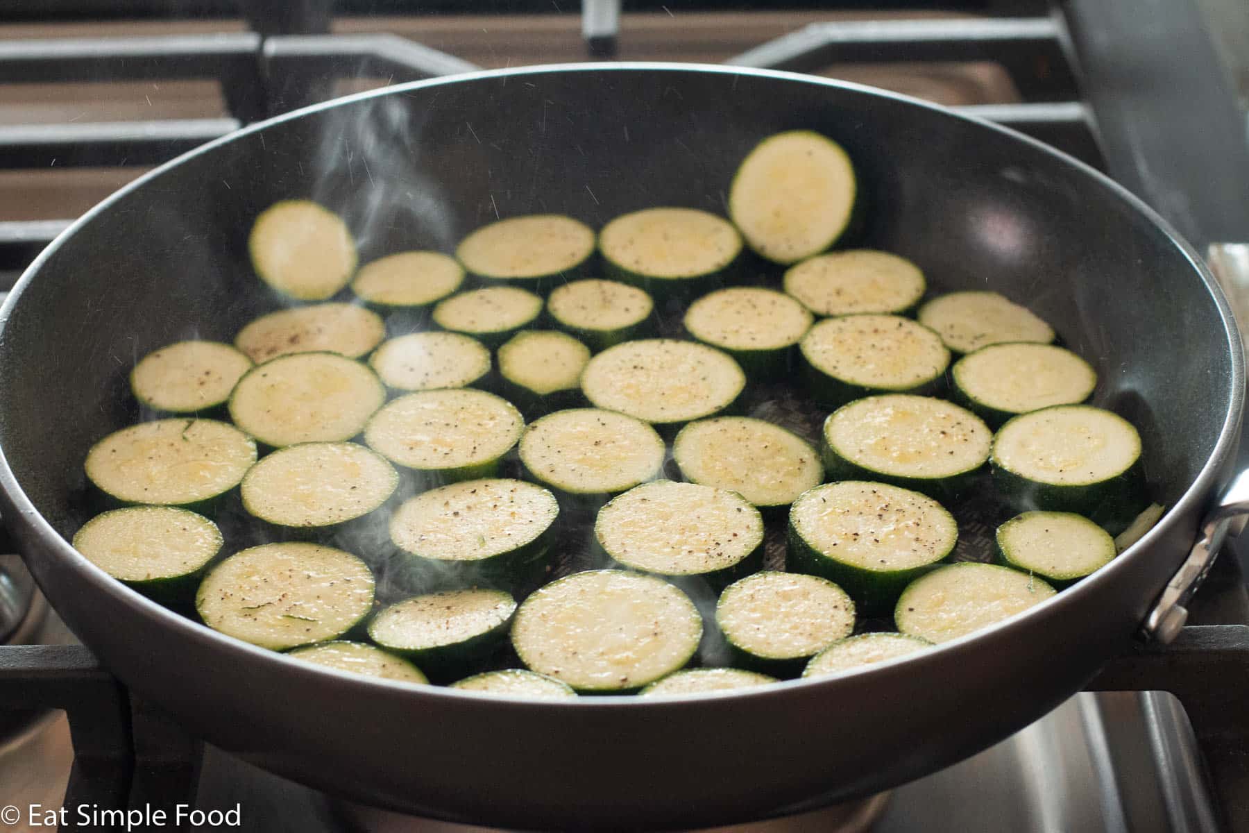 Raw half inch zucchini round slices in black pan cooking with salt and pepper on top in a single layer.