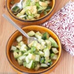 Two brown bowls with spoons of diced cucumbers and avocado salad. Green onion garnish in a black bowl on the side. White and burgundy patterned napkin on side. Close up.