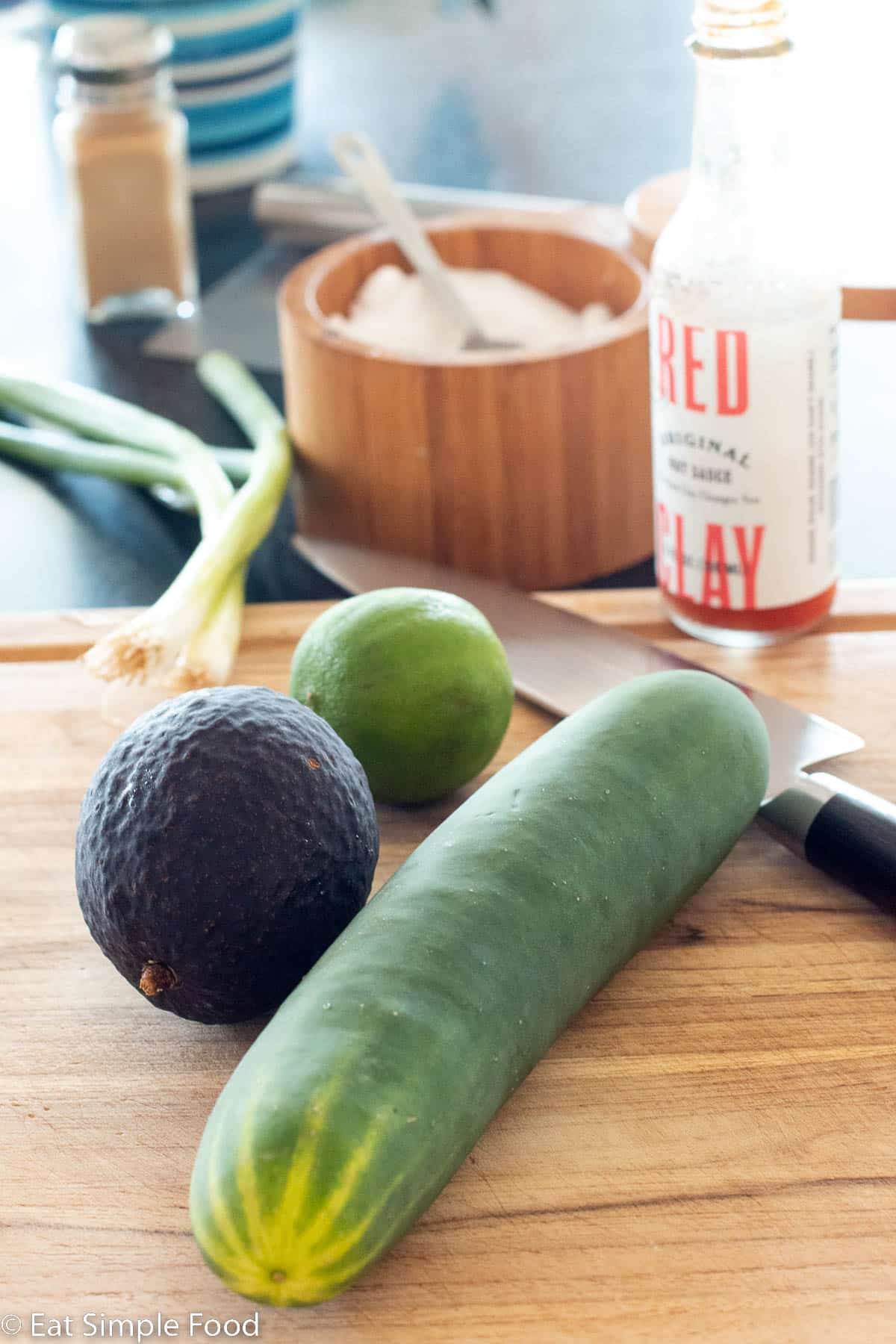 Whole cucumber, avocado, and lime with 2 green onions on a wood cutting board with a small jar of hot sauce and a container of salt. Knife and black pepper shaker in background.