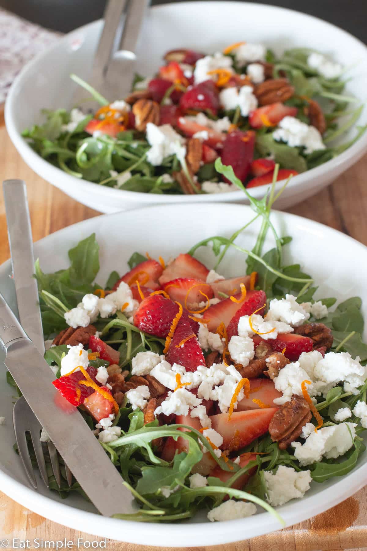 Side view of two white rimmed plates with forks and knives sticking out on a wood cutting board. Plates are filled with arugula, quartered strawberries, white goat cheese crumbles, pecans, and orange zest.