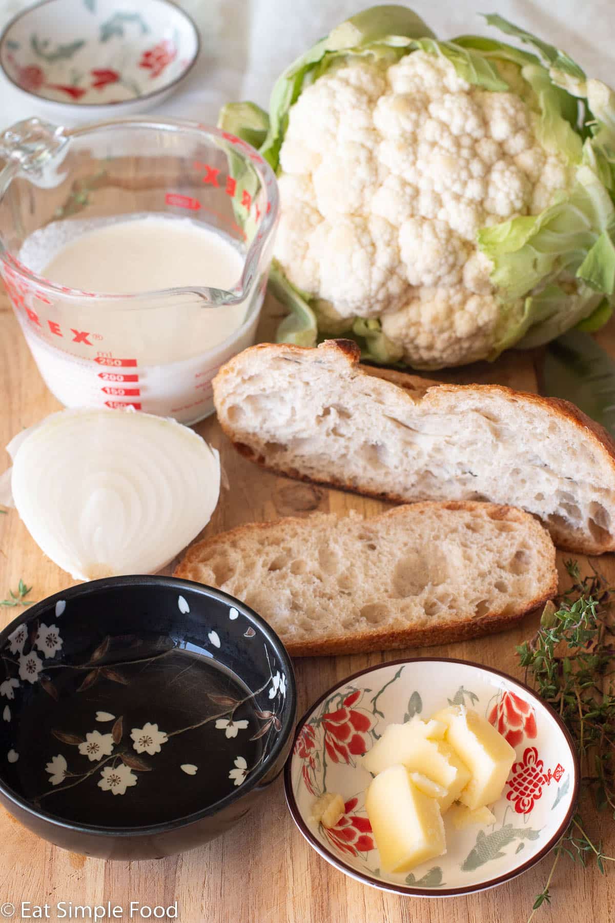 Ingredients on a wood cutting board: whole cauliflower with green leaves, 2 chunks of bread, ¾ cup heavy cream, ½ a white onions, a small bowl of butter tabs, a black bowl of liquid, and thyme sprigs on the side.