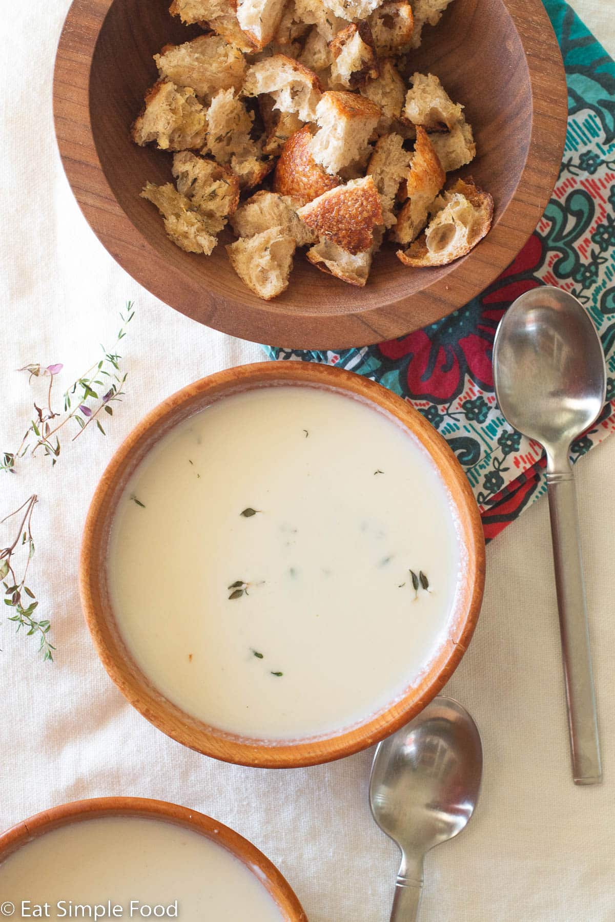 Top down view of creamy white cauliflower soup in a wood bowl with a wood bowl of croutons on the side. 2 spoons and thyme sprigs on the side.