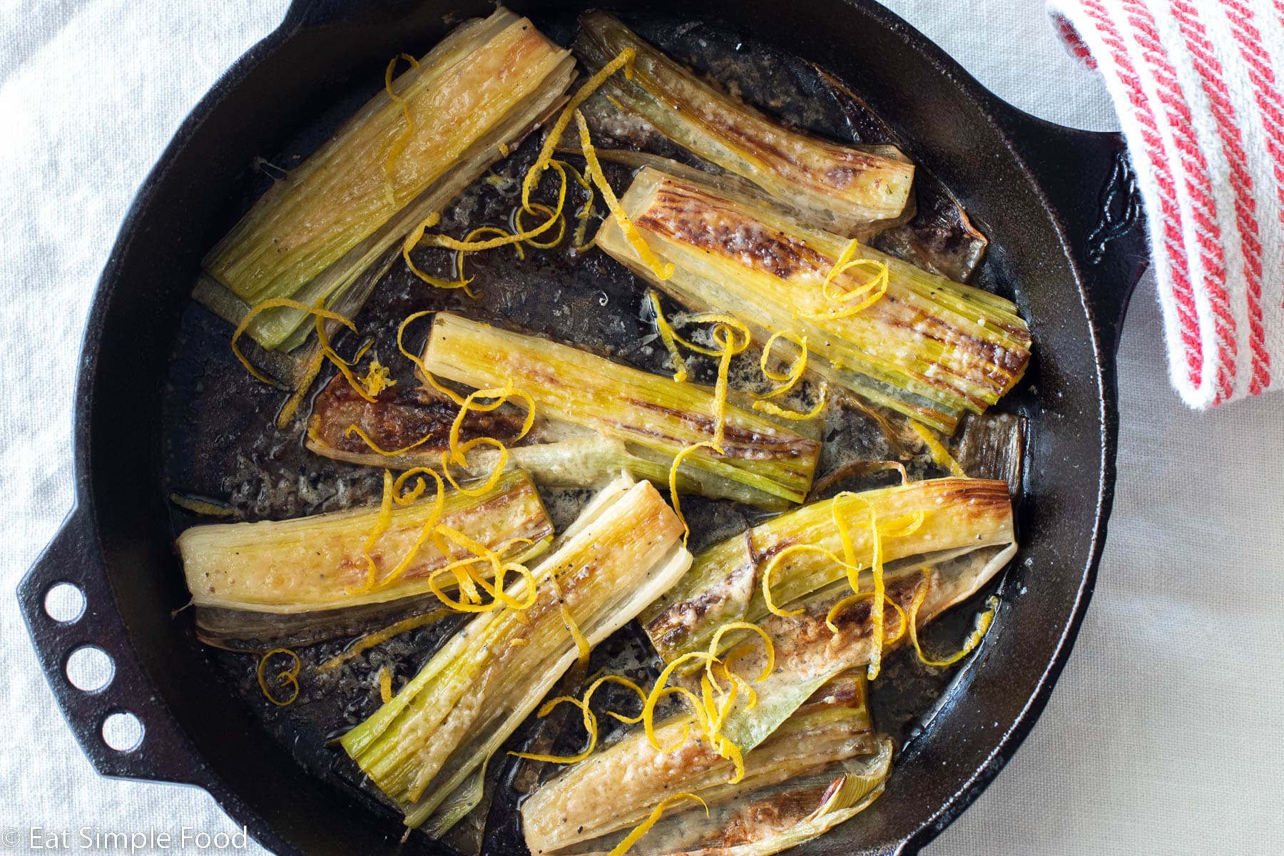 Eight seared leeks in a cast iron skillet on a white table cloth. Leeks are garnished with Parmesan cheese and lemon zest. Top down view. Red and white kitchen towel around handle of cast iron skillet.