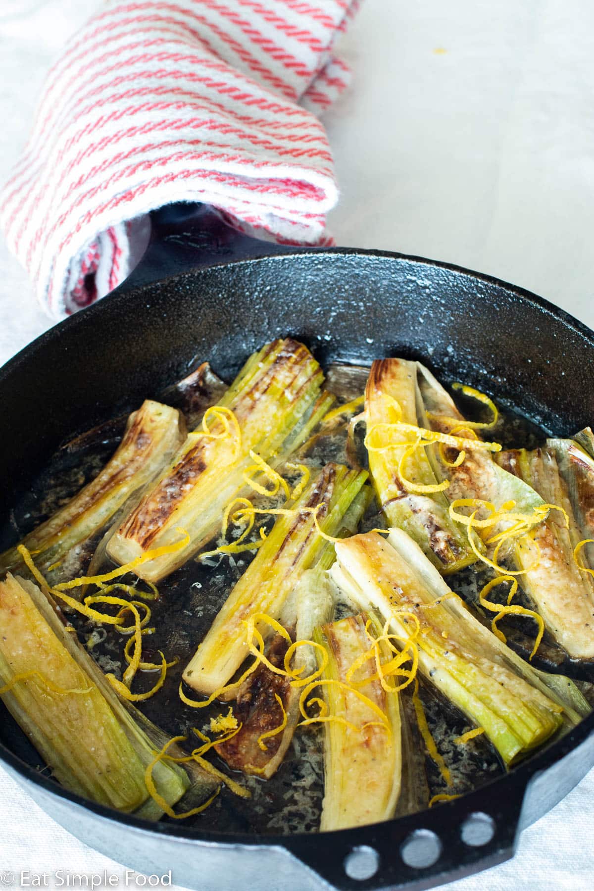 Eight seared leeks in a cast iron skillet on a white table cloth. Leeks are garnished with Parmesan cheese and lemon zest.
