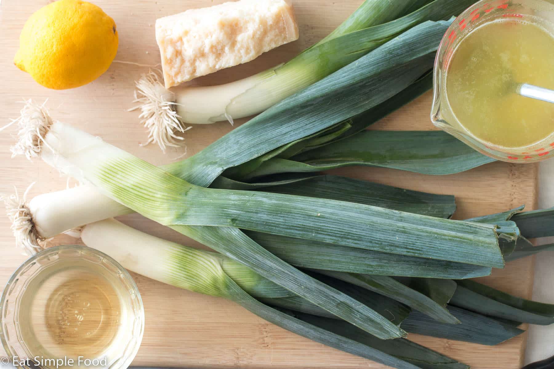 Top down view of four whole leeks, a whole lemon, a ramekin of wine, a chunk of parmesan cheese, and yellow broth in a bowl on a wood cutting board.