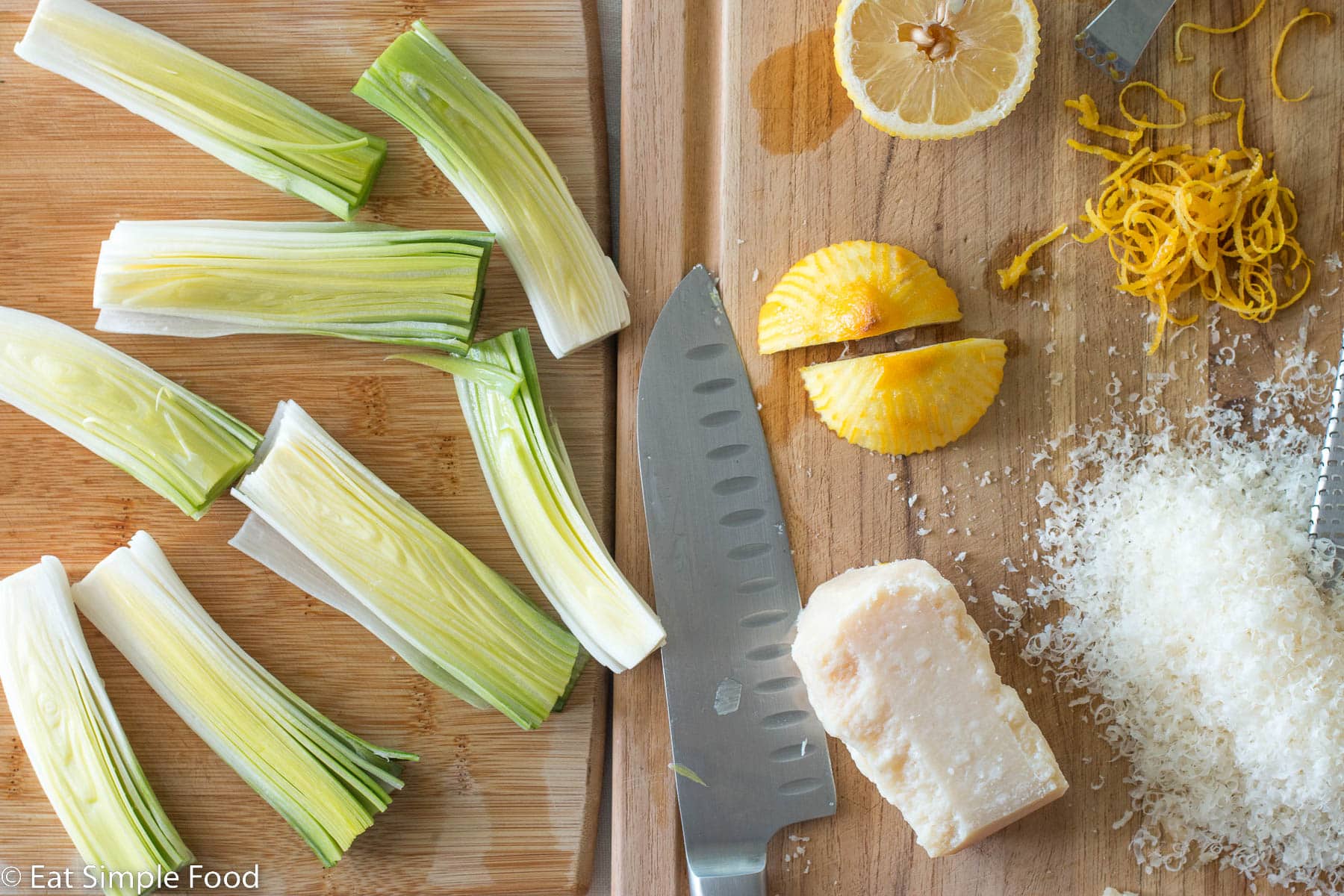 Top down view of 8 pieces of halved leeks, lemon zest, lemon halves, shredded parmesan cheese, a chunk of parmesan cheese, and a chefs knife on a wood cutting board.