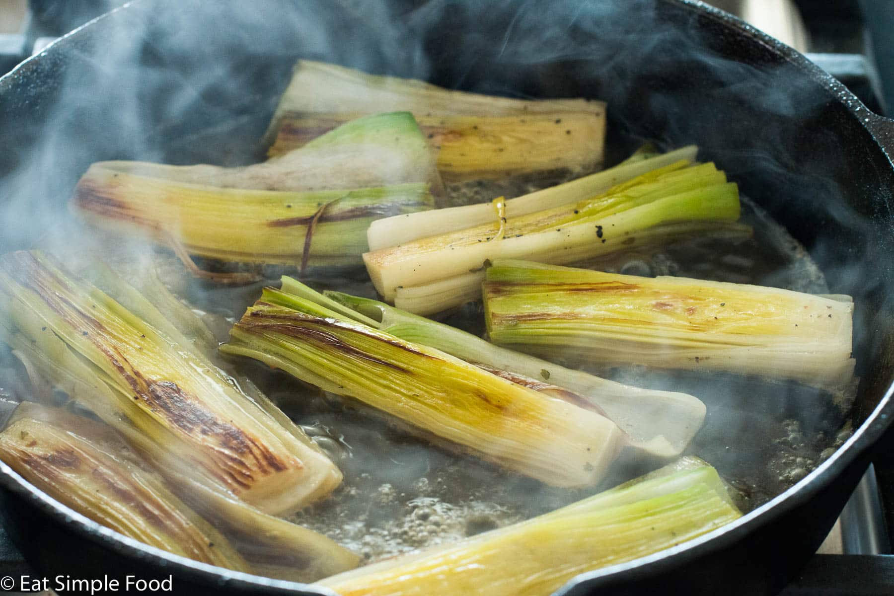 Eight leeks laved and seared side up with liquid cooking in a black cast iron skillet. Liquid is simmering and light smoke is rising.