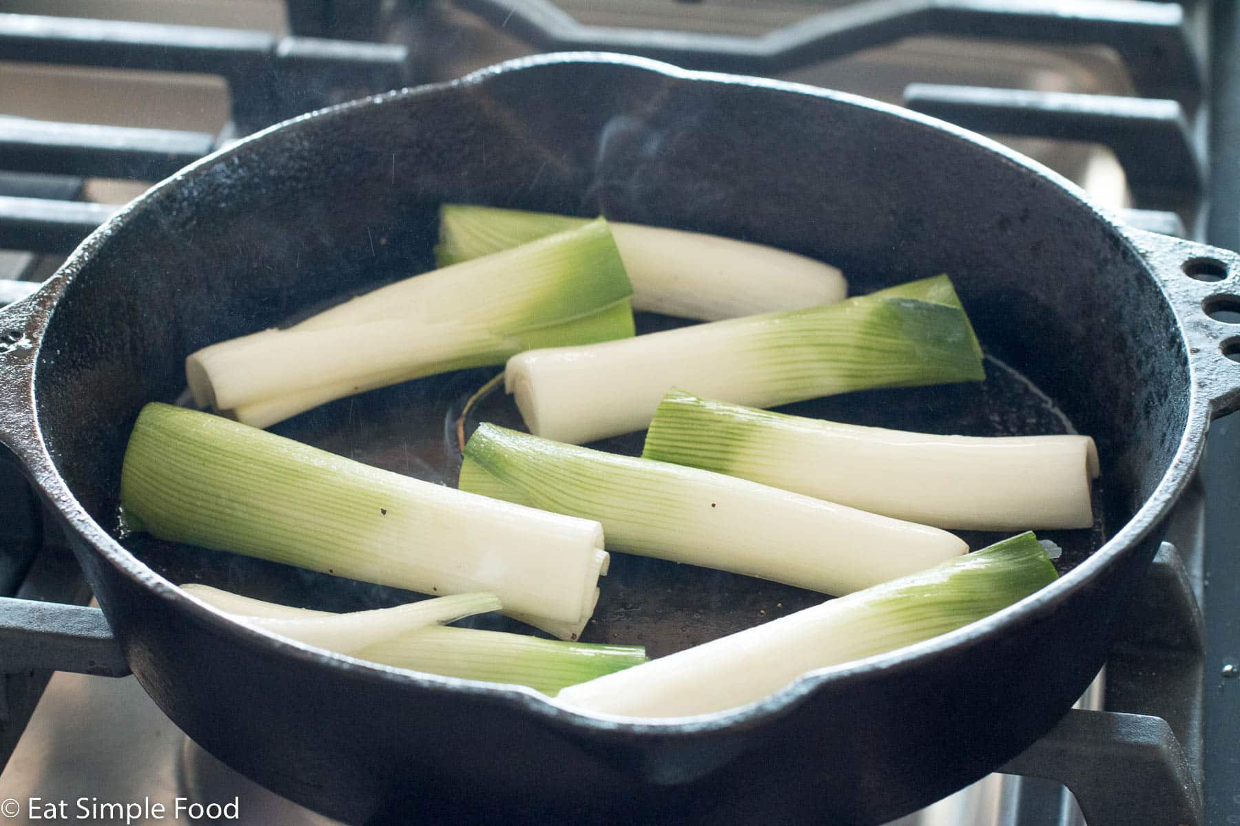 Eight halved leeks searing in a black cast iron skillet. Flat sides of leeks are facing down.