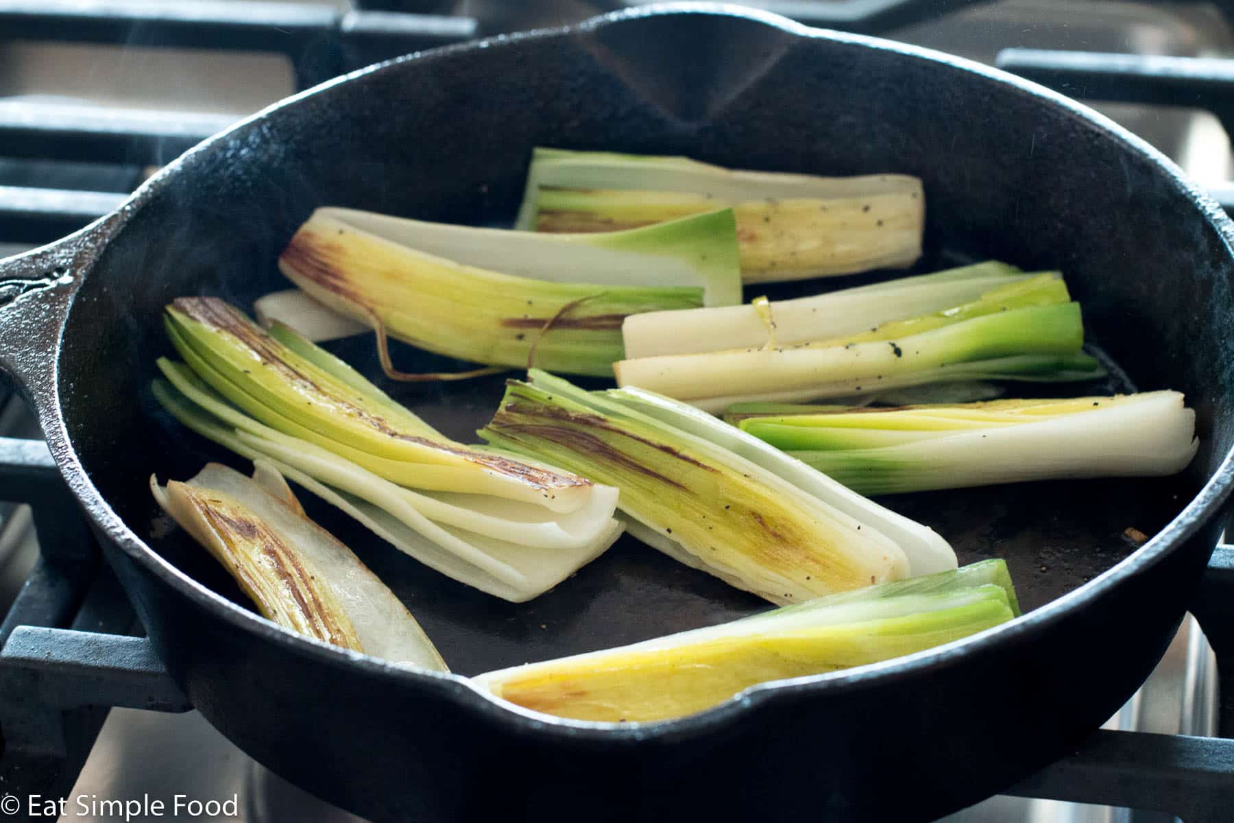 Eight halved leeks seared with the flat side facing up in a black cast iron skillet with a little smoke rising.