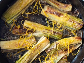 Eight seared leeks in a cast iron skillet on a white table cloth. Leeks are garnished with Parmesan cheese and lemon zest. Top down view. Red and white kitchen towel around handle of cast iron skillet. Close up.