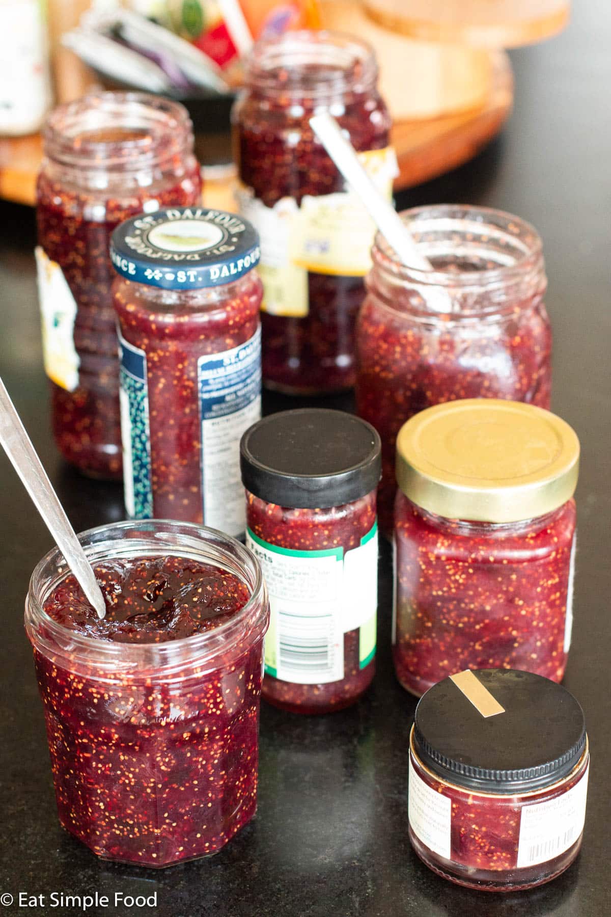 8 jars of various sizes with different lids on a black soapstone counter. 2 jars have spoons sticking out.