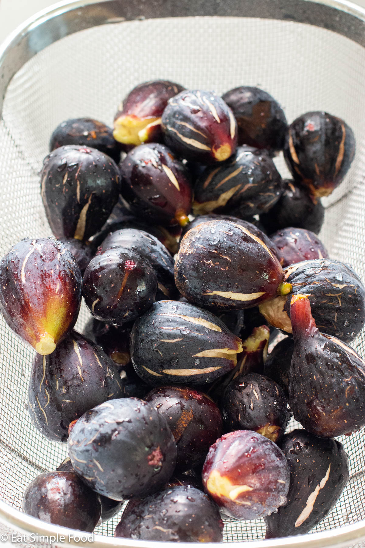 Mounds of purple brown figs piled on top of each other in a strainer. Figs are wet.