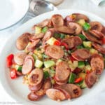 White plate of seared slices of smoked sausage on a bed of vegetables or diced red peppers, onions, and zucchini. Silverware, napkins, plates in background. Close up.