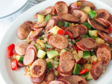 White plate of seared slices of smoked sausage on a bed of vegetables or diced red peppers, onions, and zucchini. Silverware, napkins, plates in background. Close up.