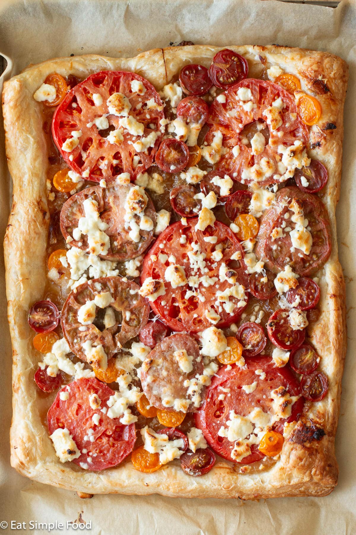 Cooked rectangle puff pastry laid on top of parchment paper. Cooked and browned ingredients on top of puff pastry layered: caramelized onions, cherry tomatoes, red and purple tomatoes, Parmesan cheese and goat cheese.