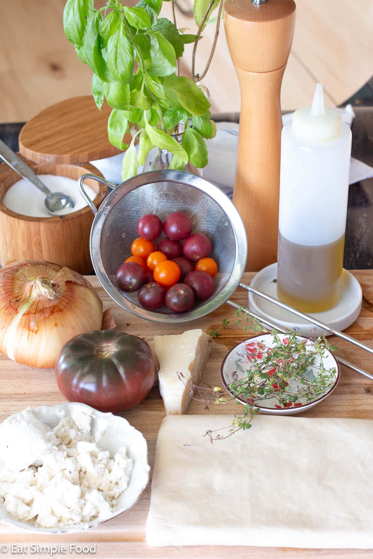 Ingredients on a wood cutting board: puff pastry rectangle, goat cheese crumbles in a white bowl, fresh thyme sprigs, chunk of Parmesan cheese, tomato, onions, small colander of cherry tomatoes, olive oil, wood jar of salt, and fresh basil sprigs in a small vase.