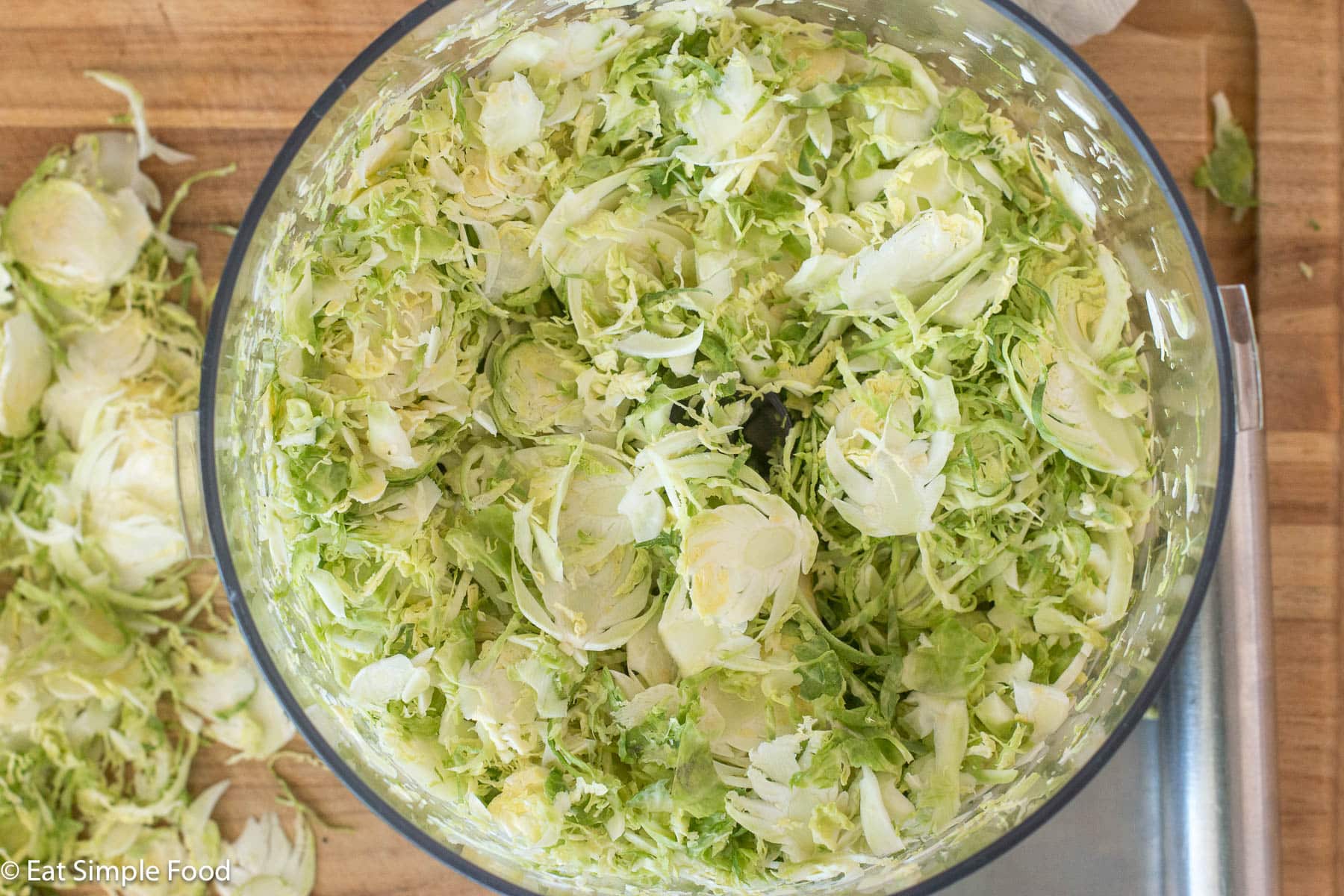 Top down view of a clear food processor full of light green shaved Brussels sprouts.