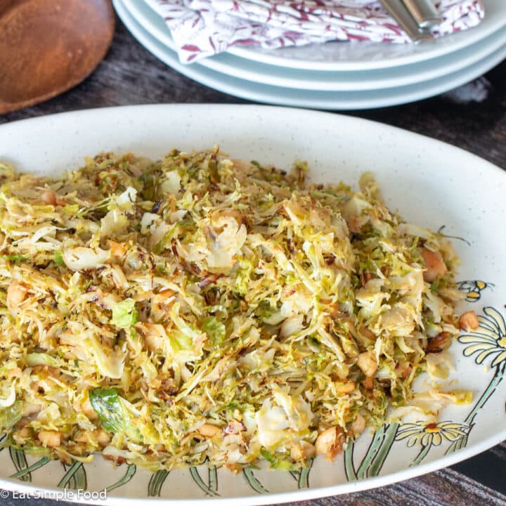White plate with yellow flowers filled with a slaw like recipe with nuts. Wood spoon and white plates in the background. Close up.
