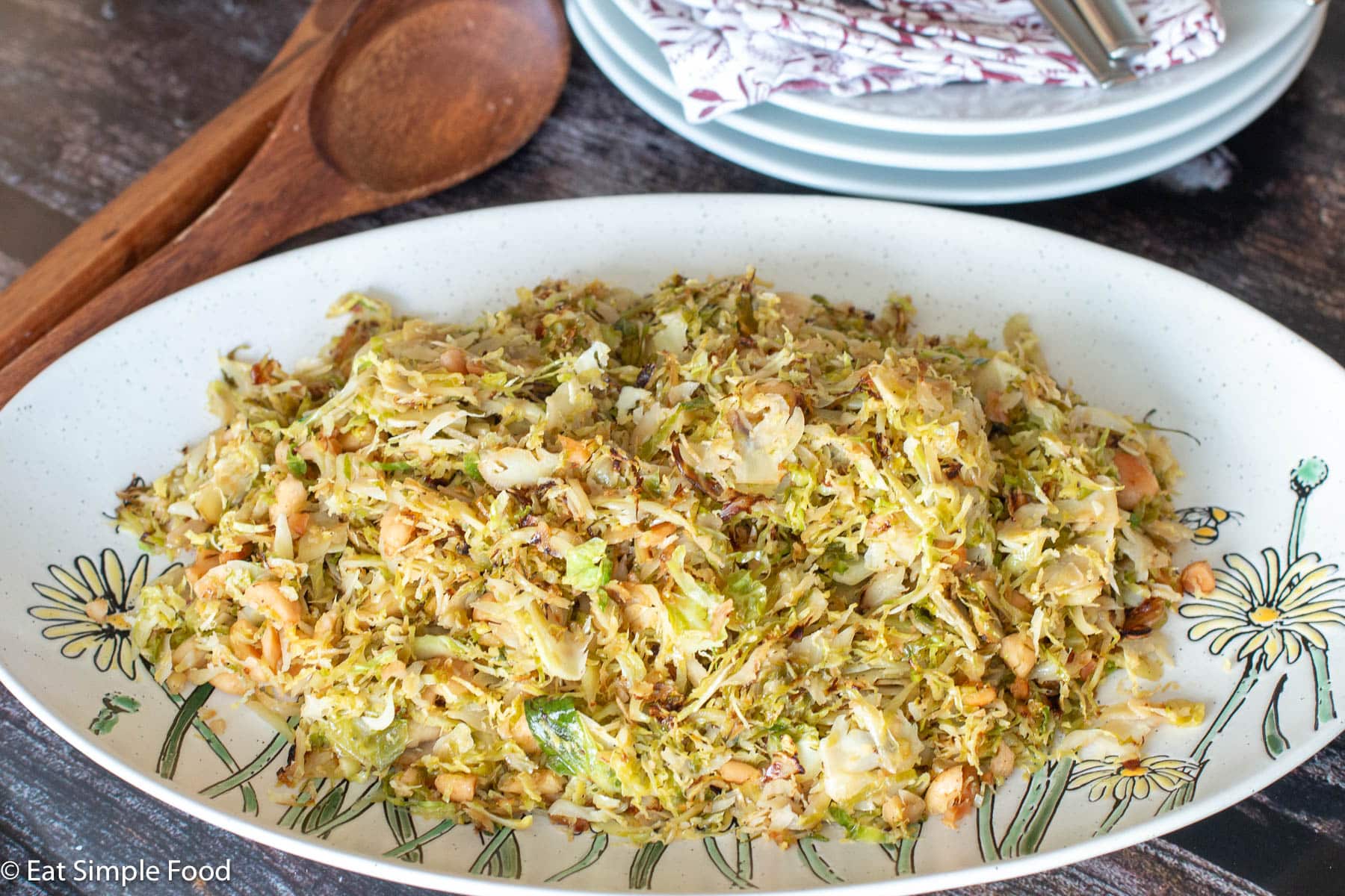 White plate with yellow flowers filled with a slaw like recipe with nuts. Wood spoon and white plates in the background.