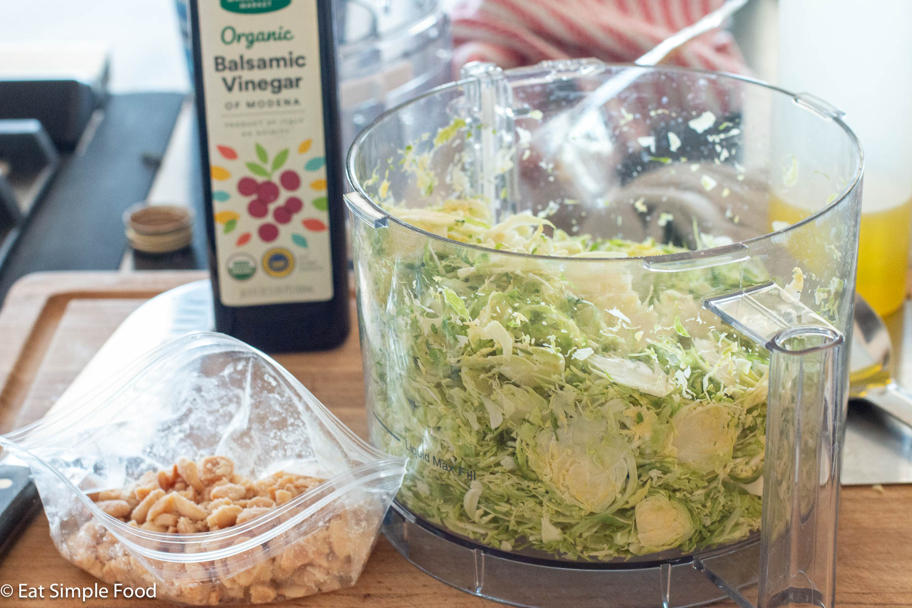 Clear food processor full of sliced Brussels sprouts with balsamic vinegar in background and a clear bag of smashed nuts on the side.