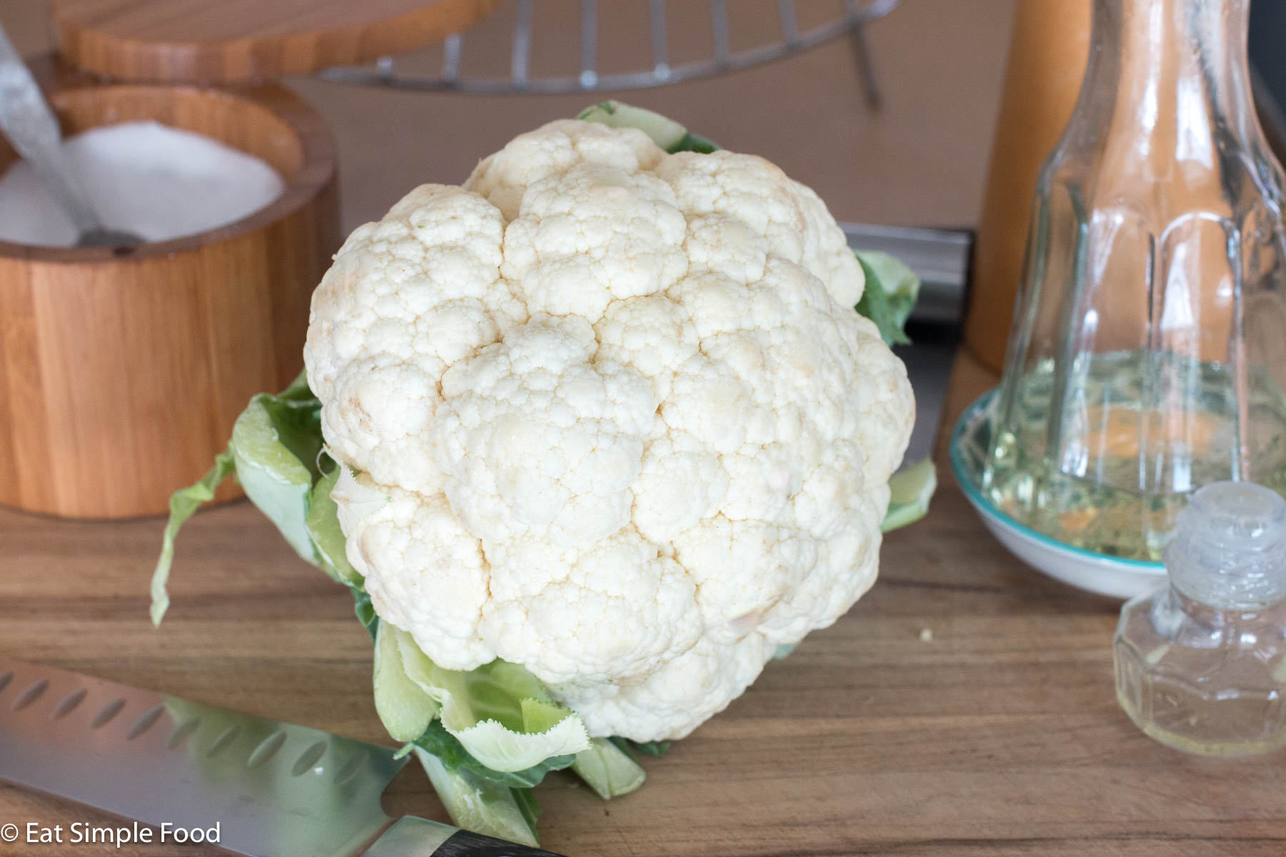Side view of a whole head of raw cauliflower on a wood cutting board with a chef's knife and salt and oil in the background.