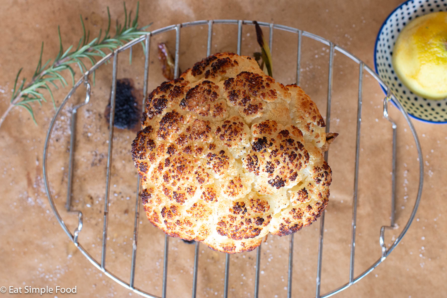 Top down view of a whole head of cauliflower cooked crispy brown on an elevated wire rack on parchment paper with rosemary sprig and lemon half on the side.
