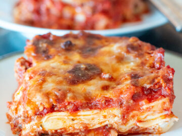 Slice of meat lasagna on a white plate with another slice in the background. Close up.
