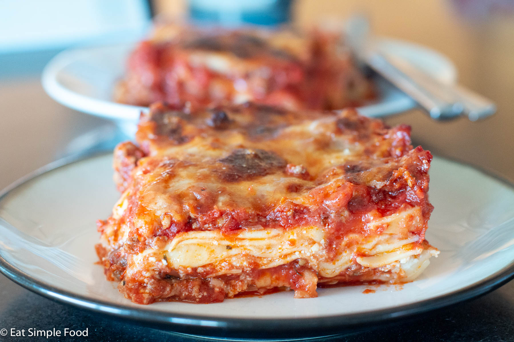 Slice of saucy meat lasagna on a white plate with another slice in the background. Close up