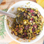 Top down view of smashed pistachios, dried cranberries, chopped rosemary in a white bowl with a spoon. Rosemary sprig and lemon half on the side. Cropped close.