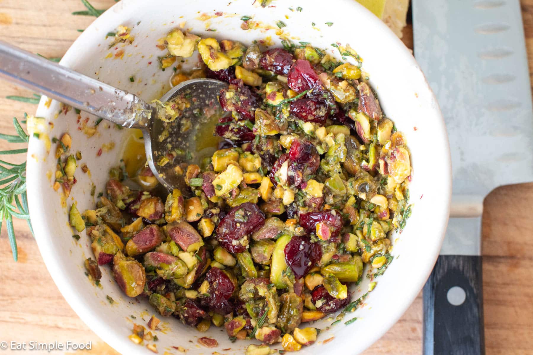 Top down view of bowl of shelled pistachios mixed with cranberries and chopped rosemary in liquid with a spoon sticking out the side.