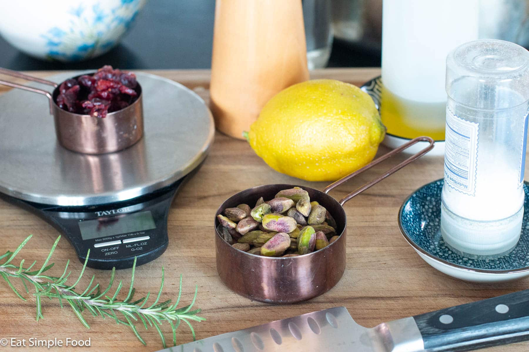 Ingredients on a wood cutting board: ½ cup of shelled pistachios, ¼ cup of cranberries, salt shaker, 1 whole lemon, oil in container, sprig of rosemary, and a chefs knife.