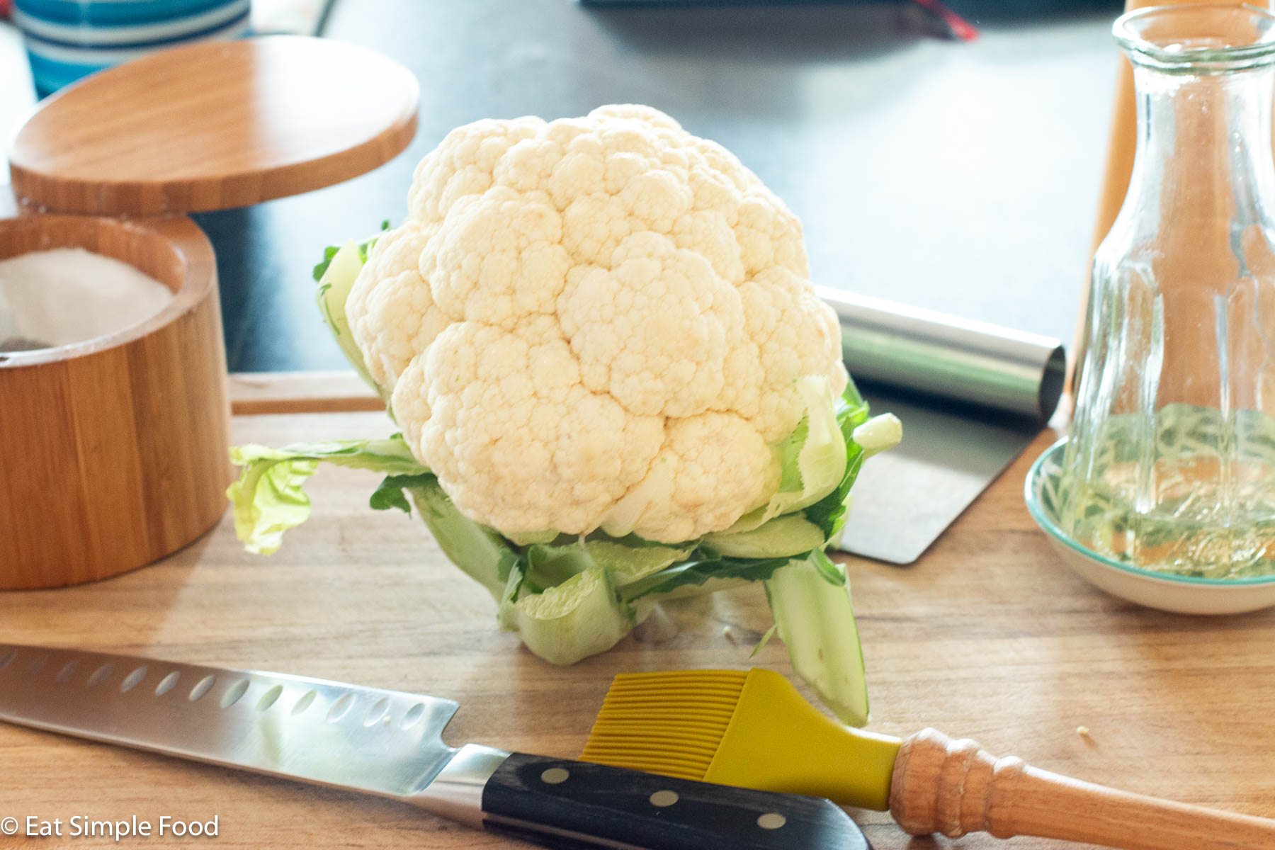 Cauliflower head on wood cutting board with a bottle of oil, container of salt, silicone baster, and chefs knife.