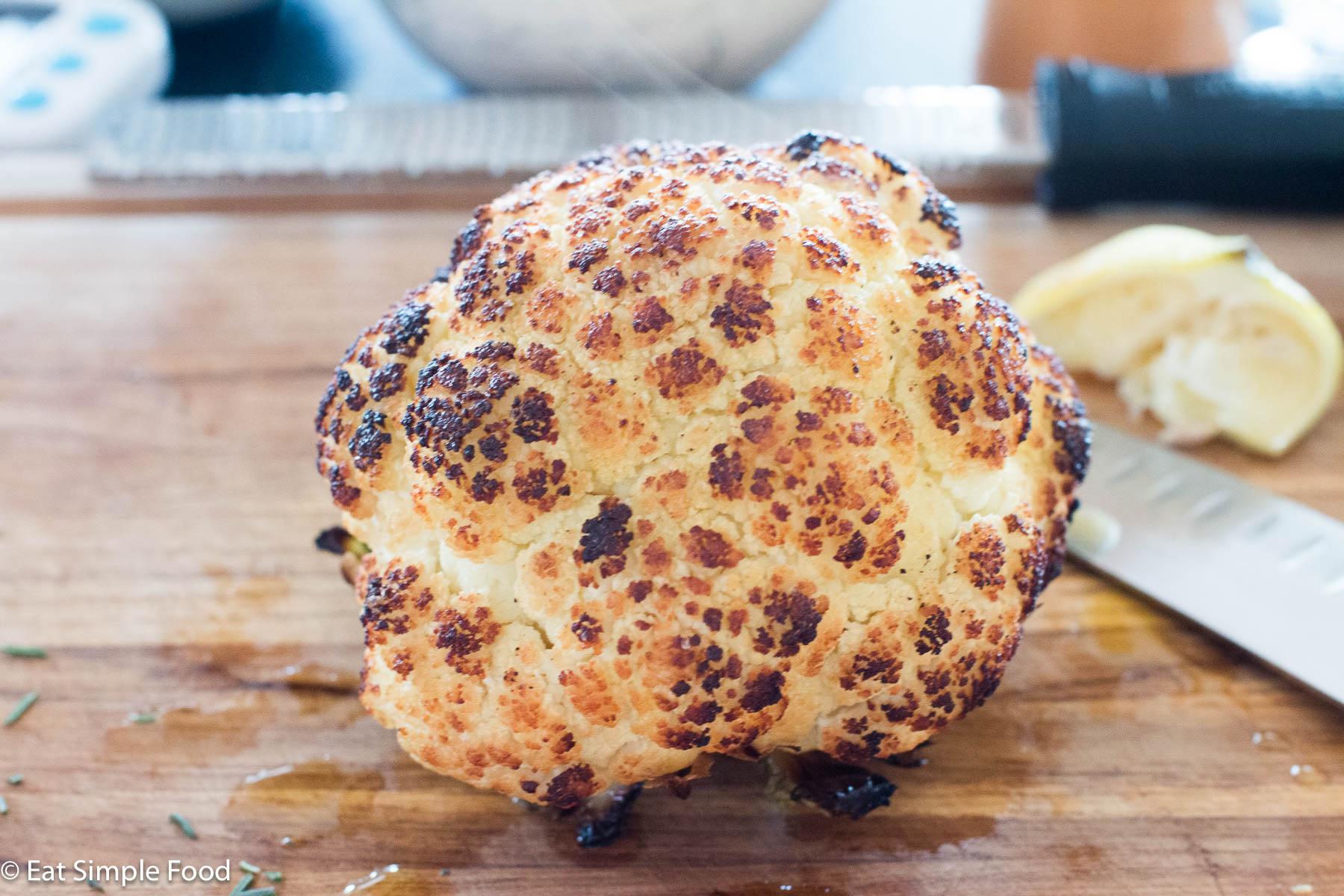 Whole browned roasted cauliflower on a wood cutting board with a lemon half and zester in the background.