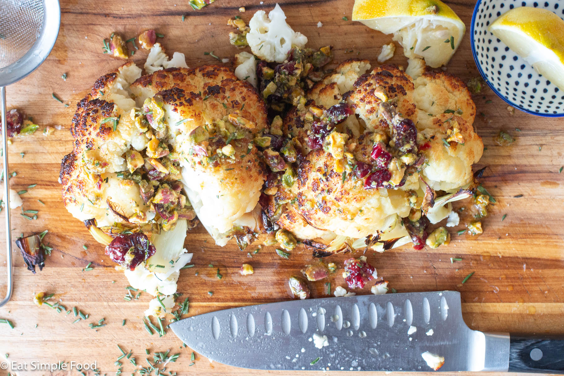 Two halves of roasted browned caulflower flat side down on a wood cutting board with a chefs knife and lemon half. Cranberries, chopped rosemary and pistachios layered over the top.