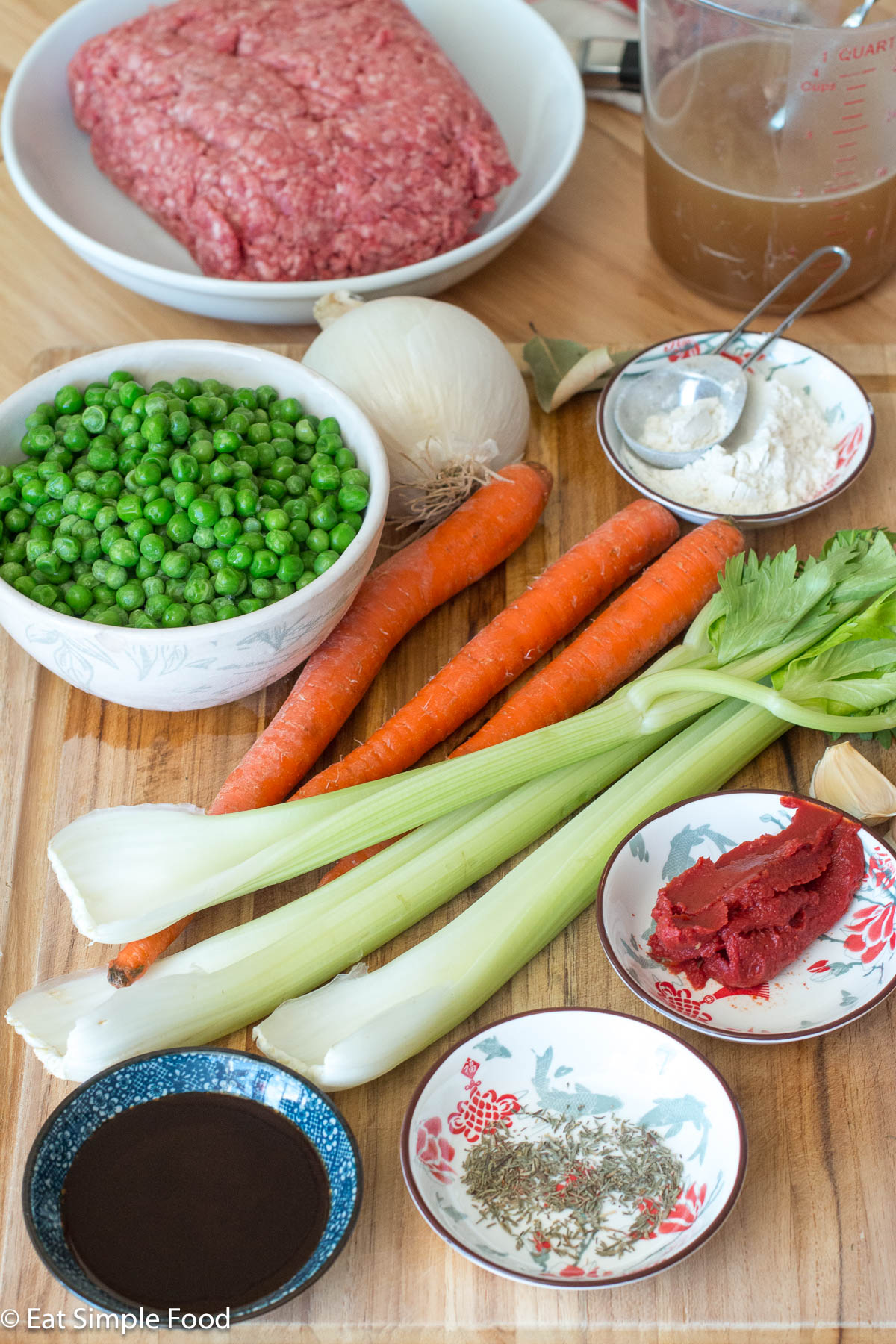 Ingredients on a wood cutting board: 3 carrots, 3 celery stalks, bowl of green peas, white onion, garlic cloves, tomato paste, herbs and a plate of raw ground beef in the background.